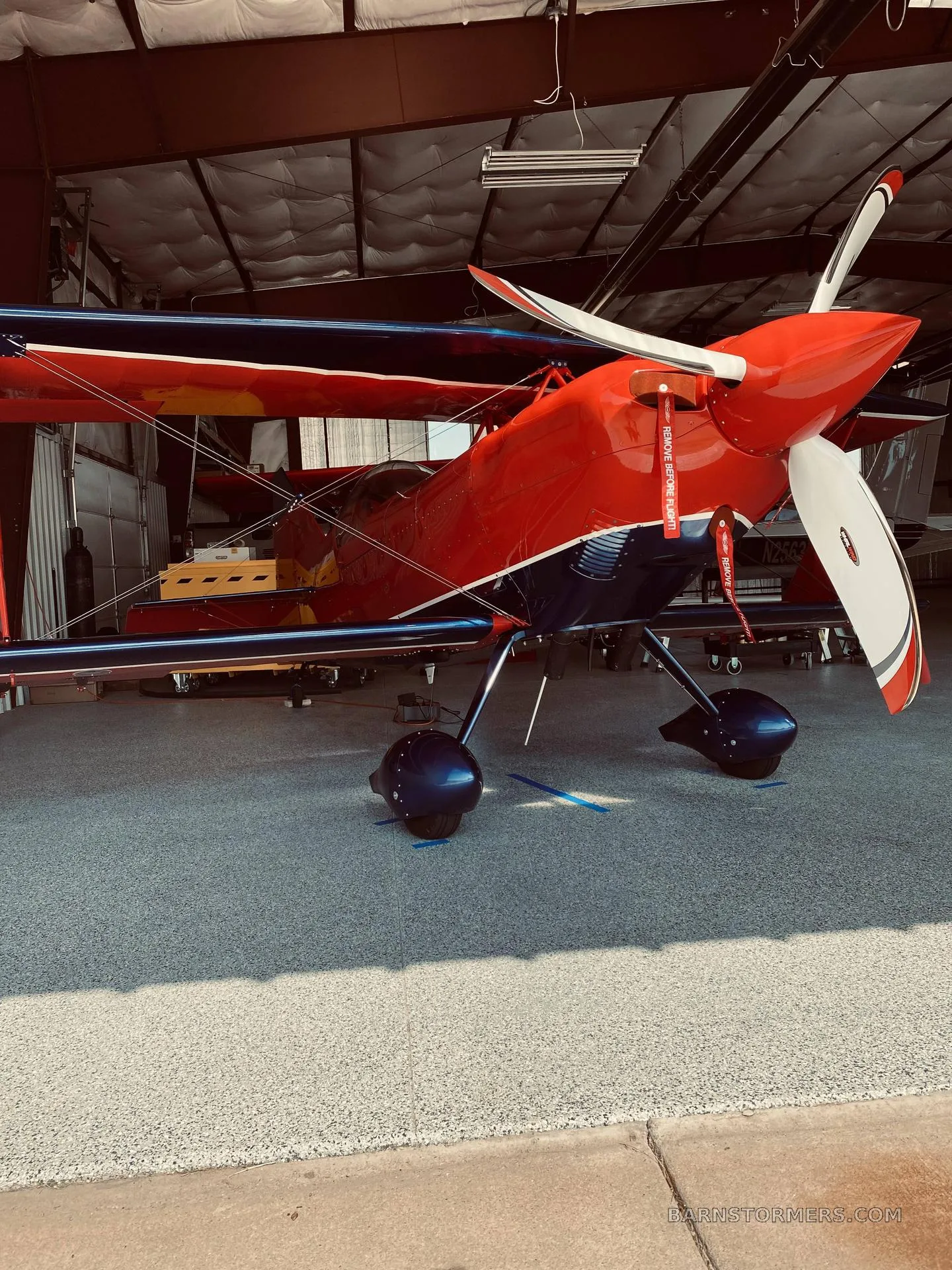 A red and blue biplane with "REMOVE BEFORE FLIGHT" tags is parked inside a hangar.