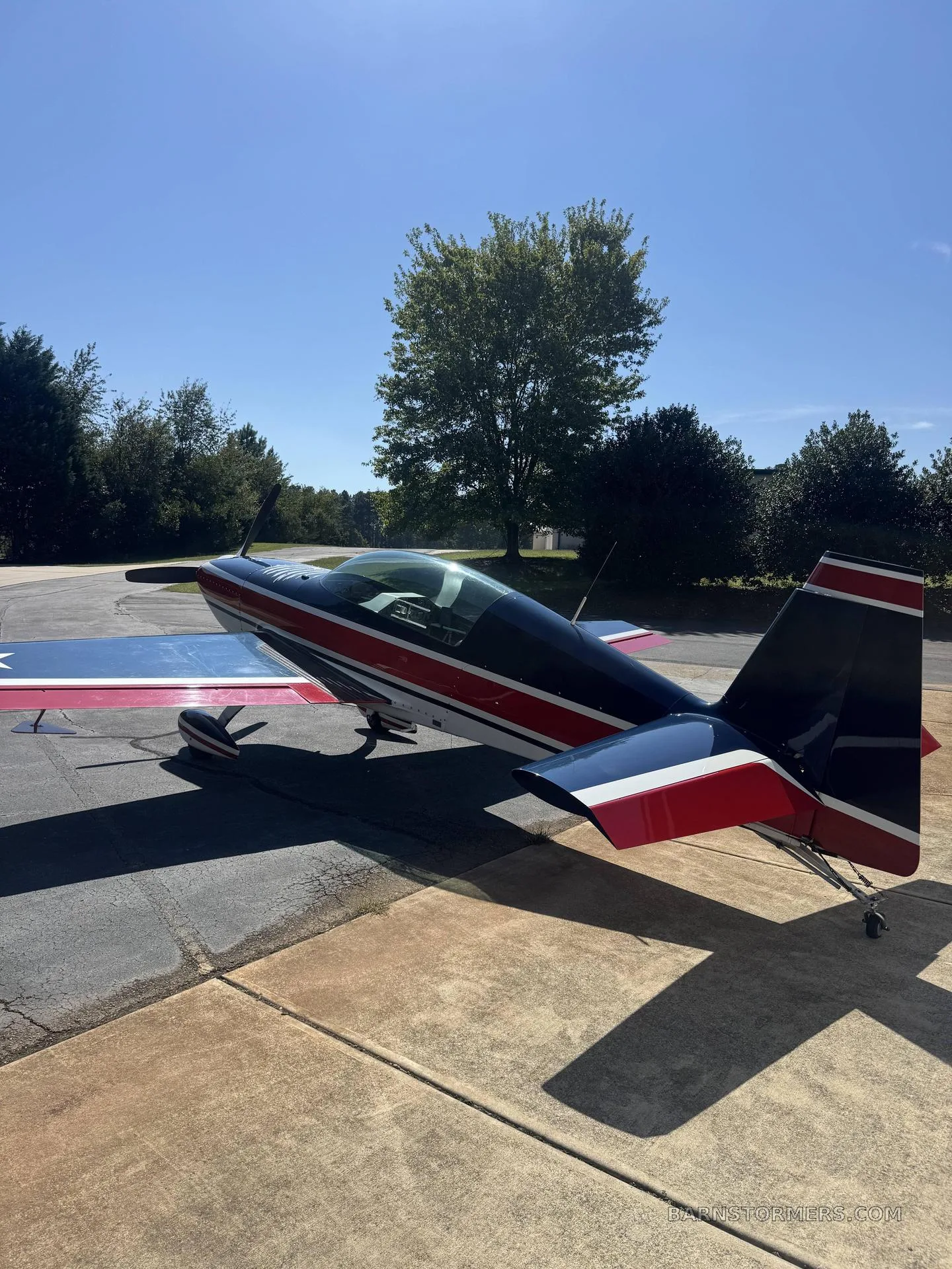 A dark blue, red, and white aerobatic aircraft with stars on its wings, parked on a concrete pad under a clear blue sky.