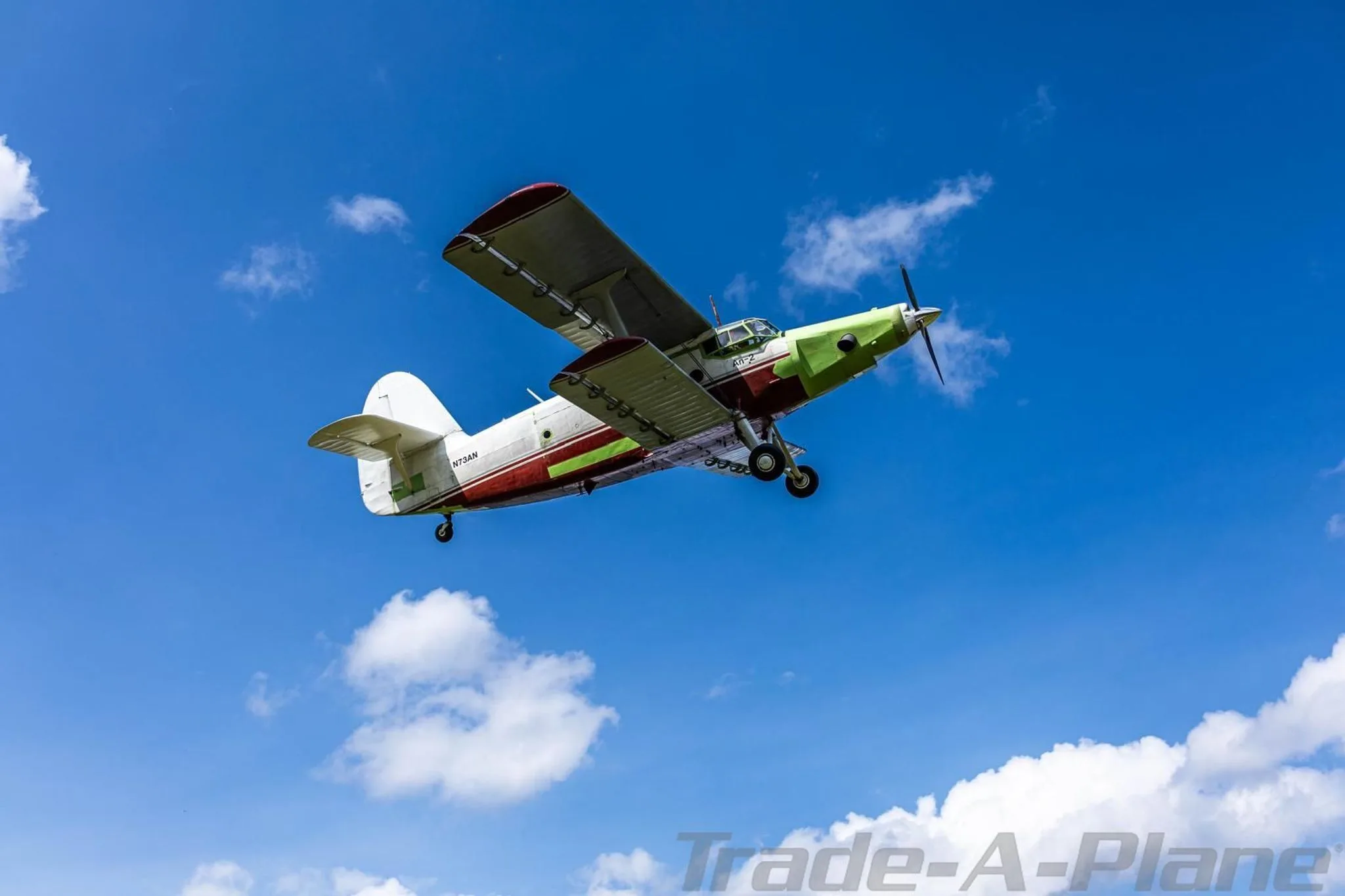 An Antonov An-2 biplane, painted white, red, and green, flying against a bright blue sky with scattered clouds.