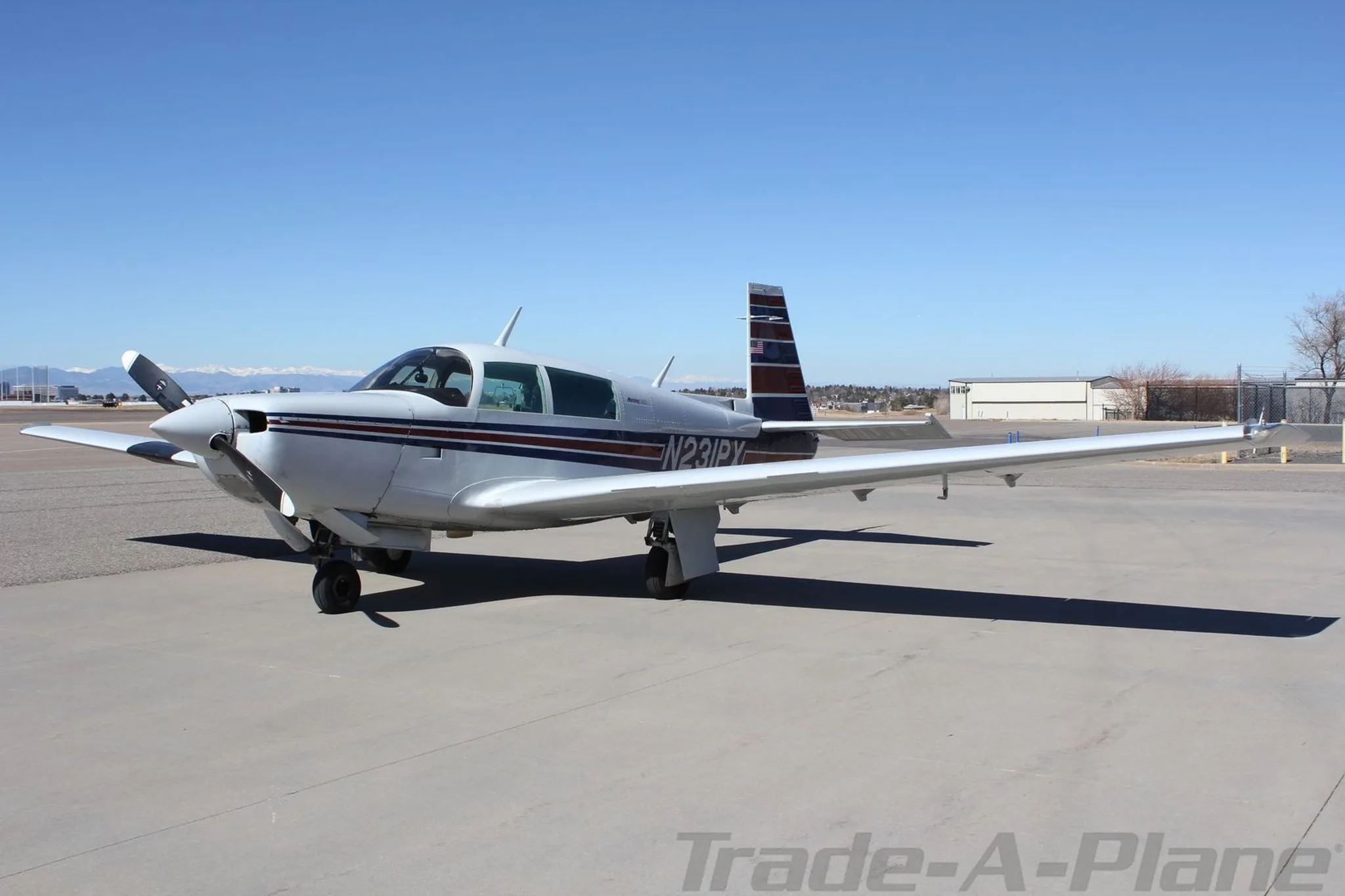 A silver single-engine aircraft with blue and red stripes on the tail and fuselage, parked on an asphalt ramp under a clear blue sky.