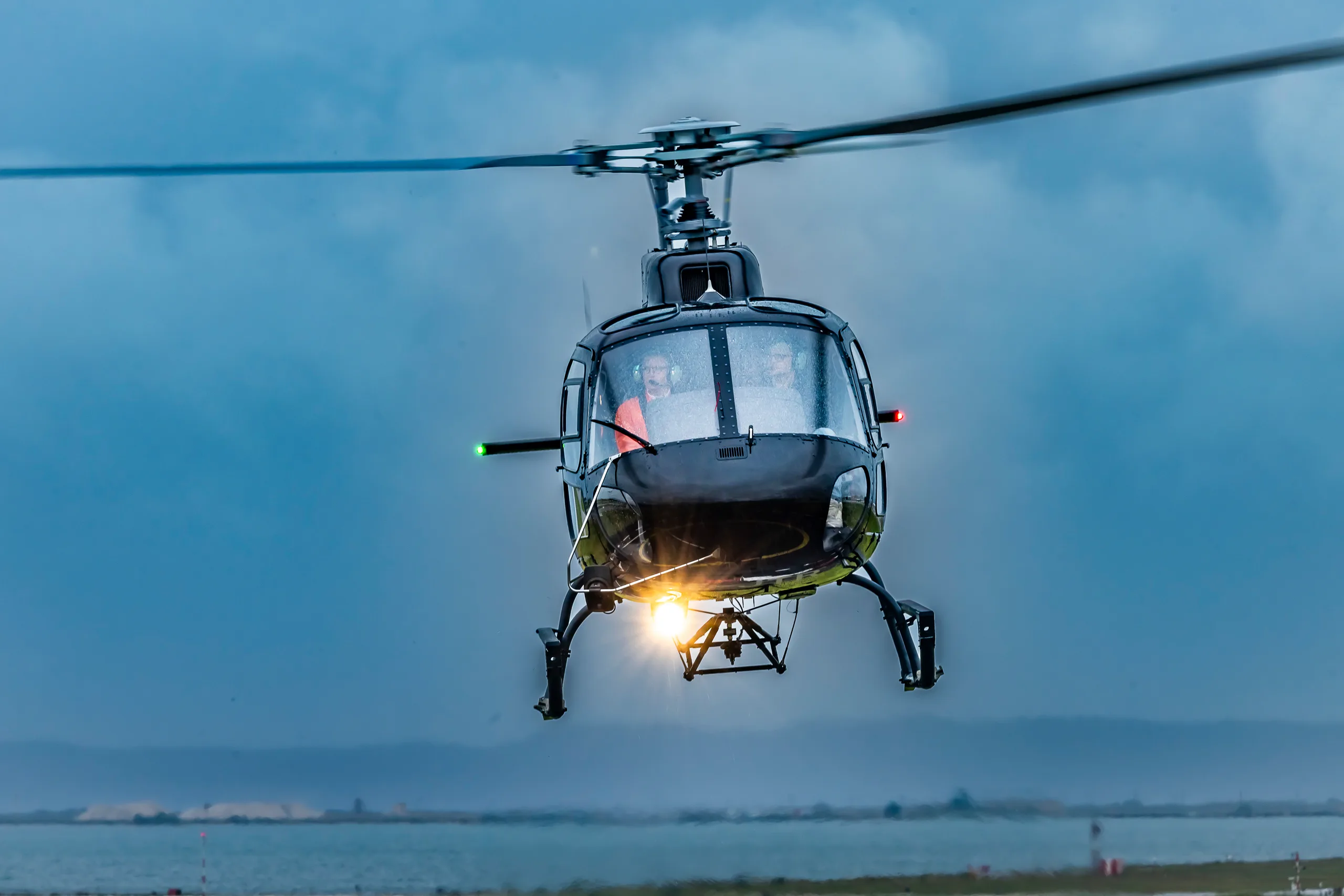 A black helicopter with its landing light on hovers over water with land in the background under a cloudy sky.
