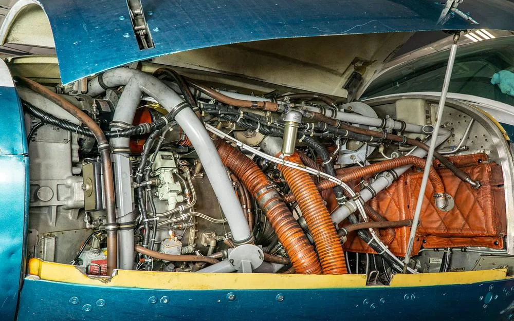The open engine cowling of a blue aircraft reveals complex wiring and pipes.
