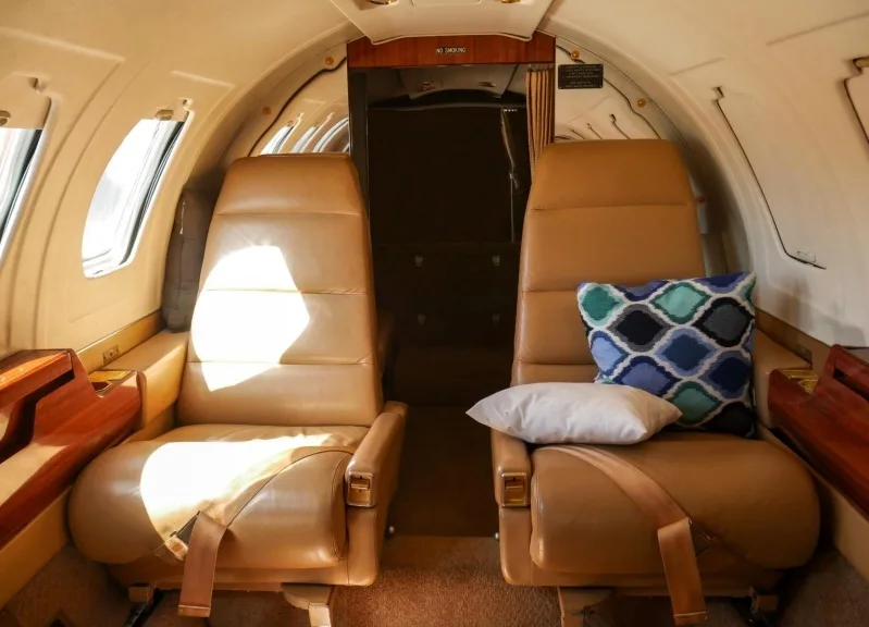 Two tan leather passenger seats in the cabin of a private jet, with a patterned pillow on one seat.