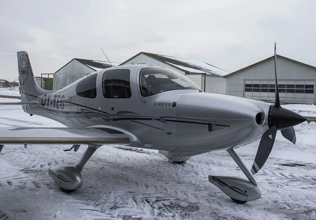 A silver Cirrus SR22 Turbo aircraft with black accents, registration OY-TEG, parked on a snowy tarmac in front of hangars.