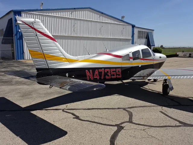 A silver, black, red, and yellow single-engine propeller aircraft, N47359, parked on an asphalt ramp next to a hangar.