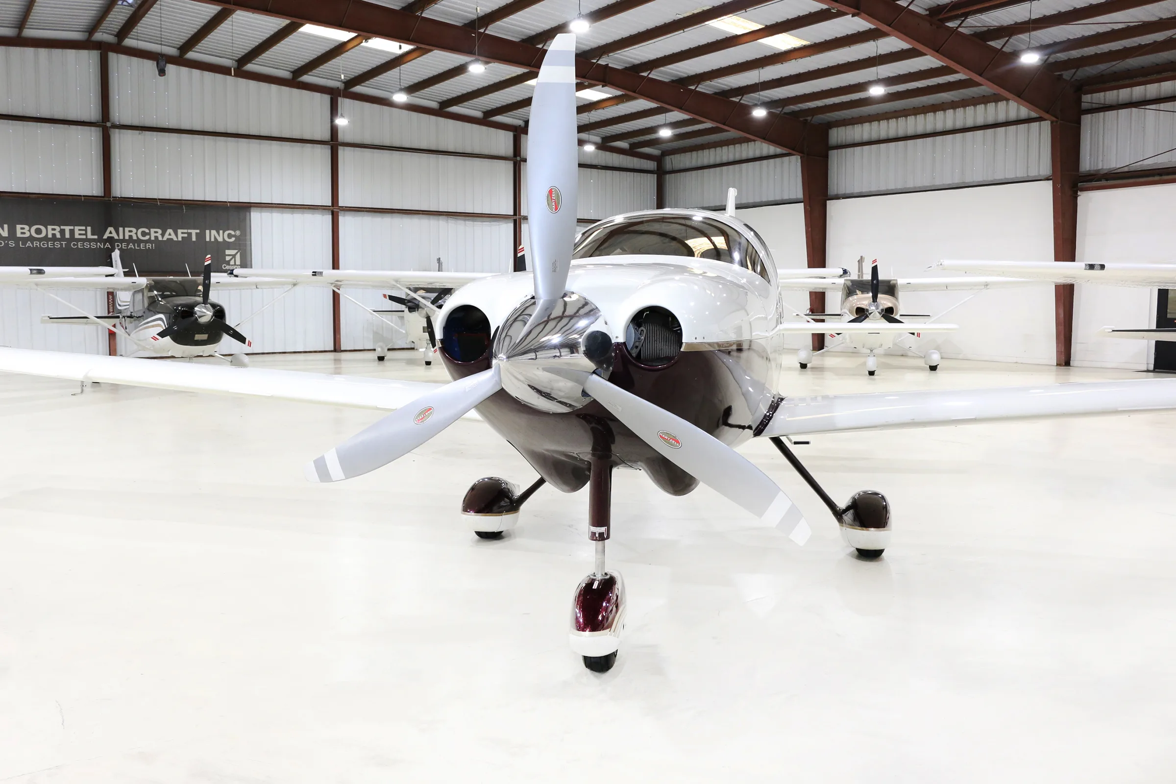A white and brown single-engine propeller aircraft is parked in a hangar, viewed from the front.