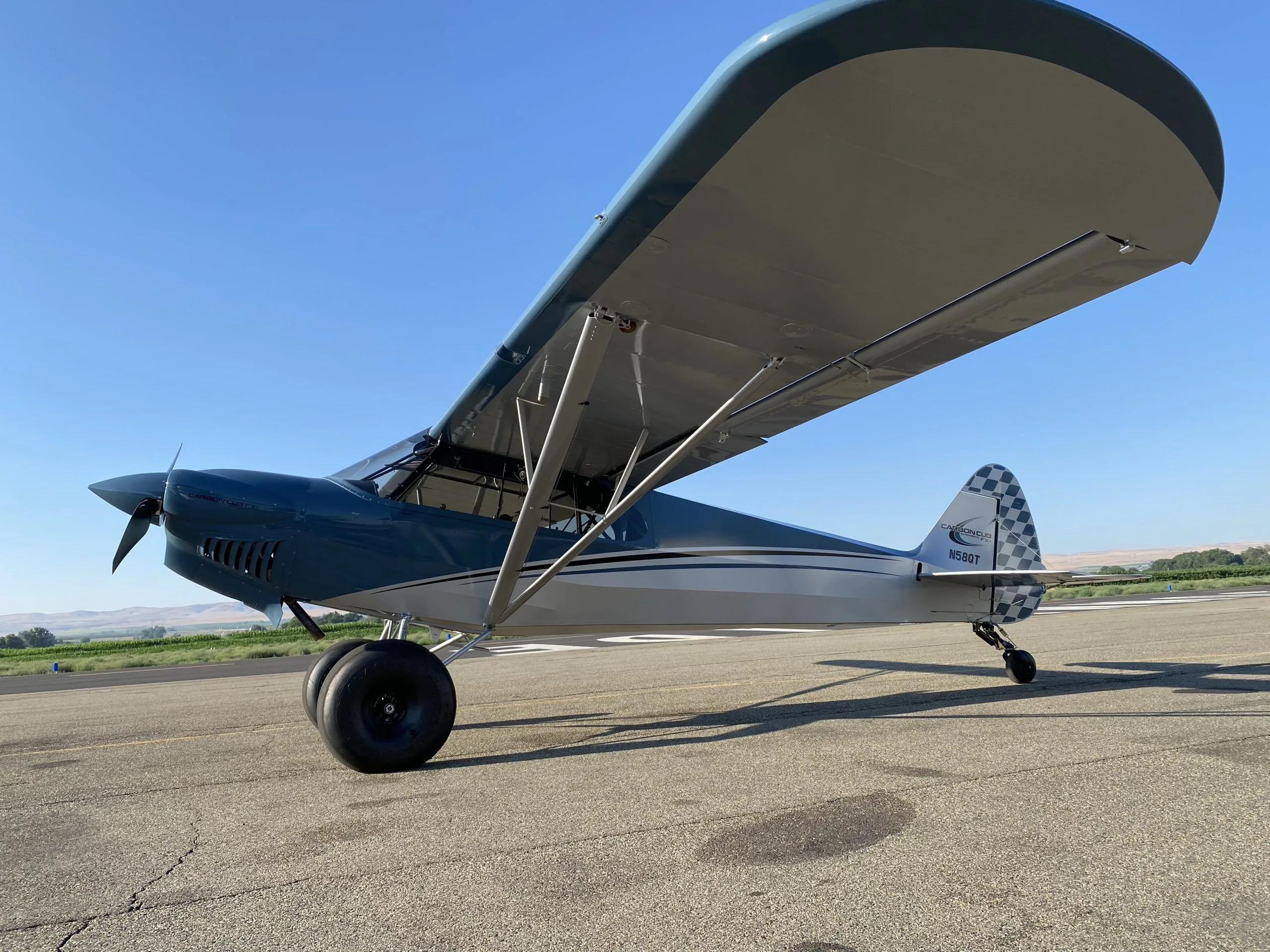 A blue and grey Carbon Cub FX-3 aircraft with registration N58QT parked on a tarmac, viewed from a low angle.