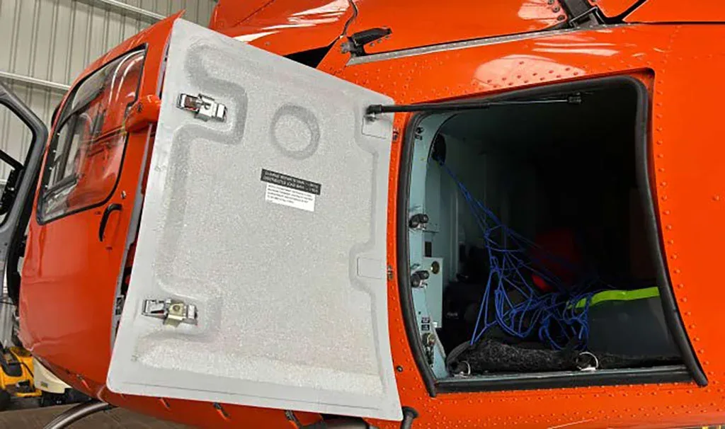 A close-up view of an open cargo door on the side of a bright orange helicopter, revealing a storage compartment with blue netting.