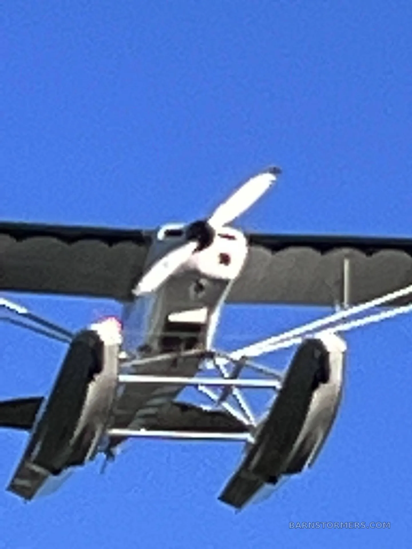 A white seaplane with floats and a propeller is seen from below, flying against a clear blue sky.