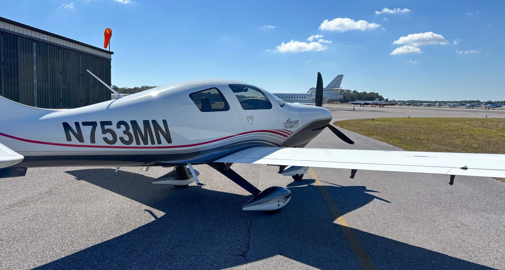 Side view of the Lancair Columbia 400 N753MN parked on the tarmac with a hangar and other aircraft in the background.