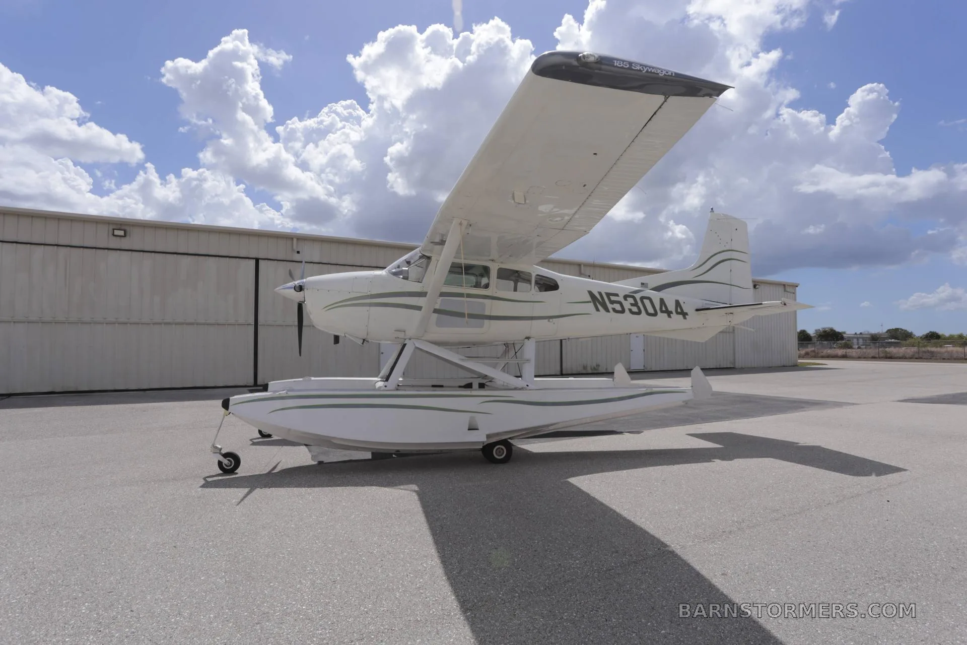 A white Cessna 180 Skywagon seaplane with green stripes and registration N53044 is parked on an airfield under a cloudy sky.