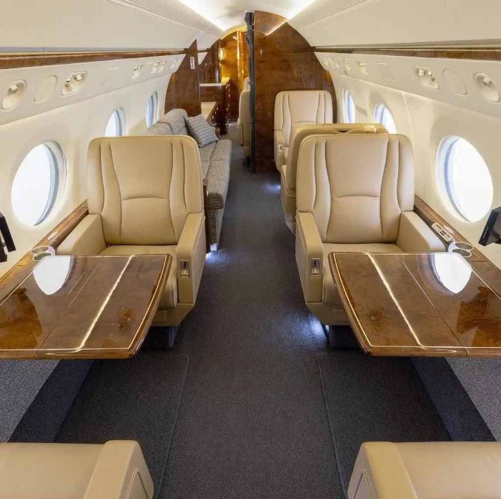 The spacious cabin interior of a business jet featuring tan leather seats and glossy wood tables.