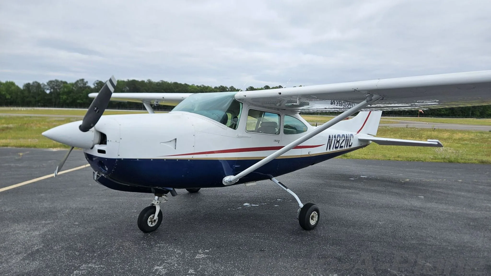 A white and blue Cessna 182 with red and gold stripes is parked on an asphalt tarmac under a cloudy sky.