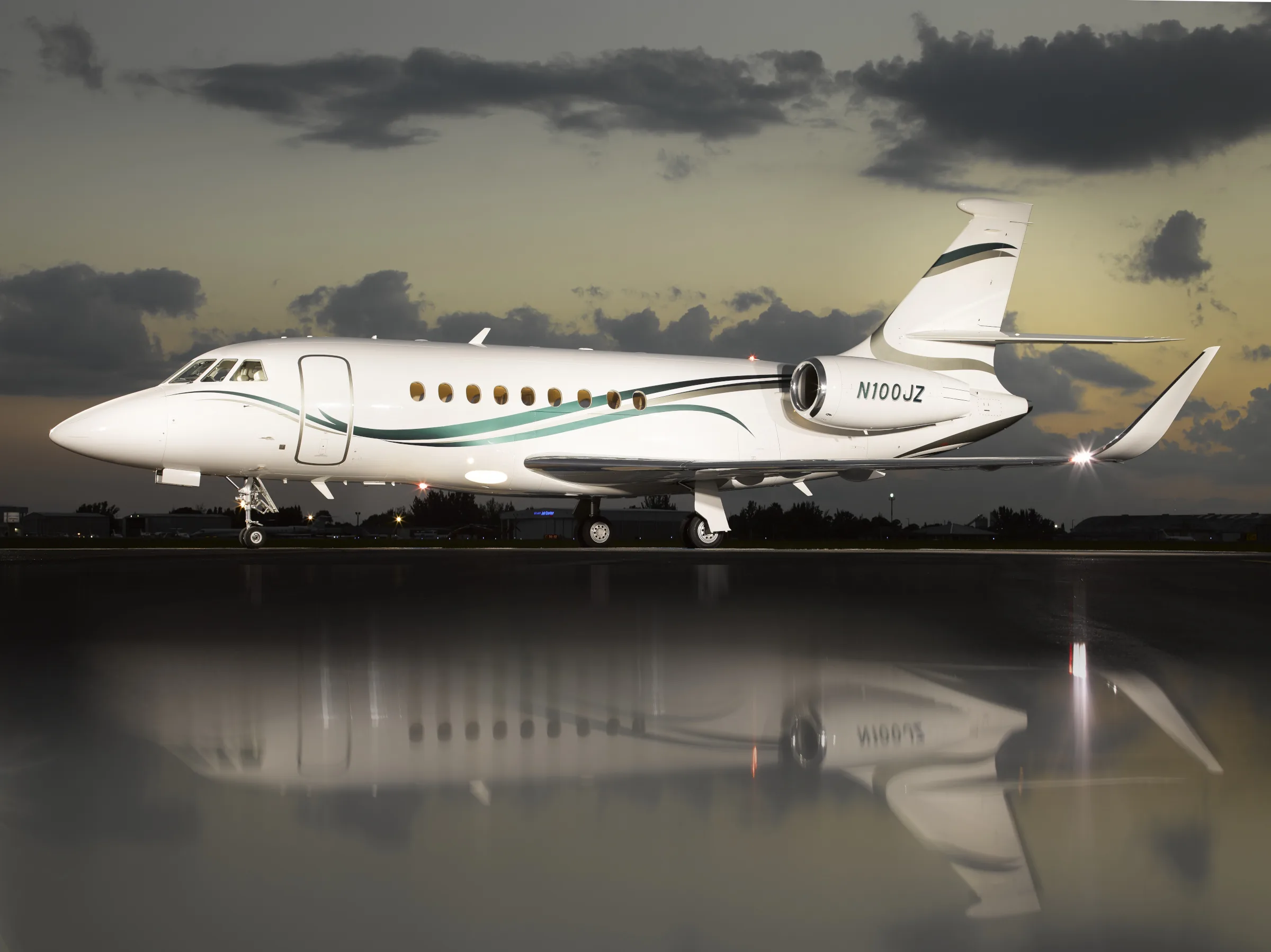 A white Dassault Falcon 2000LXS business jet with green and grey livery parked on a wet tarmac at sunset.