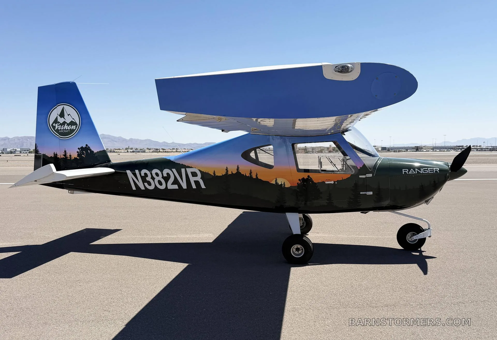 A green and blue 'Ranger' light-sport aircraft with a forest and sunset paint scheme, seen from the side-right on an airfield.