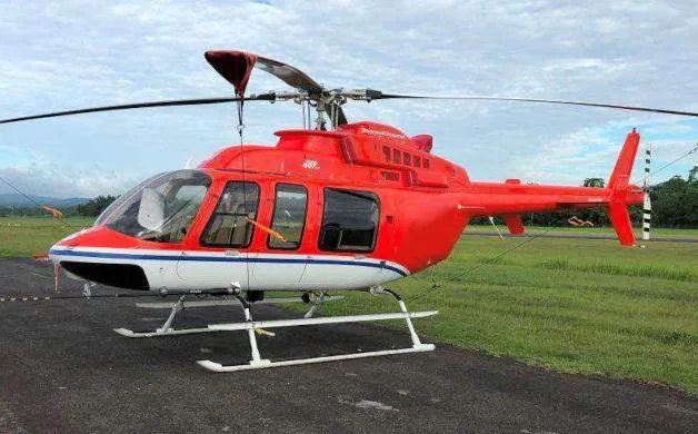 A red and white Bell 206 helicopter with blue stripes is parked on an asphalt pad under a cloudy sky.