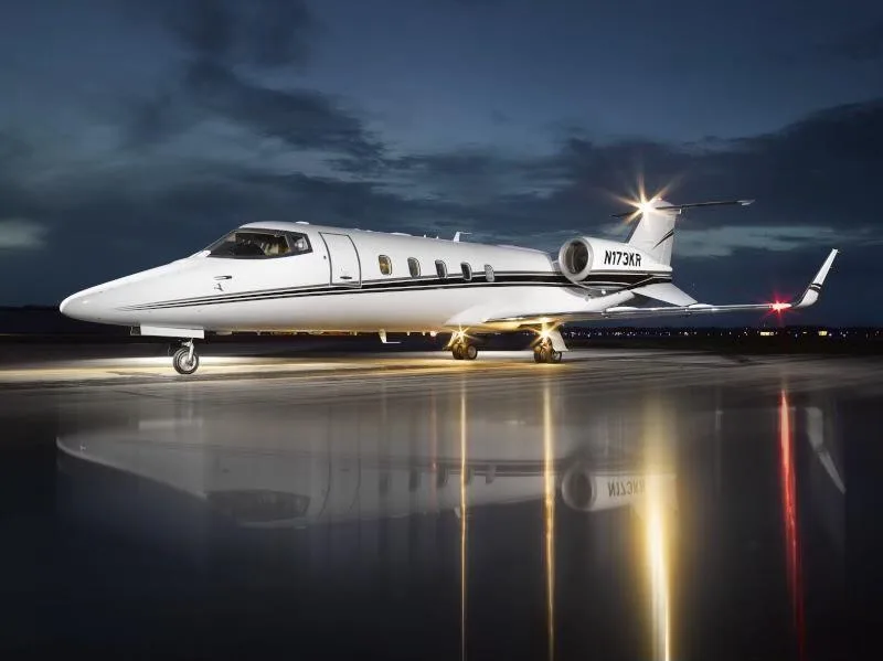 A white Learjet with black stripes, tail number N173KR, parked on a wet tarmac at night with landing lights on.