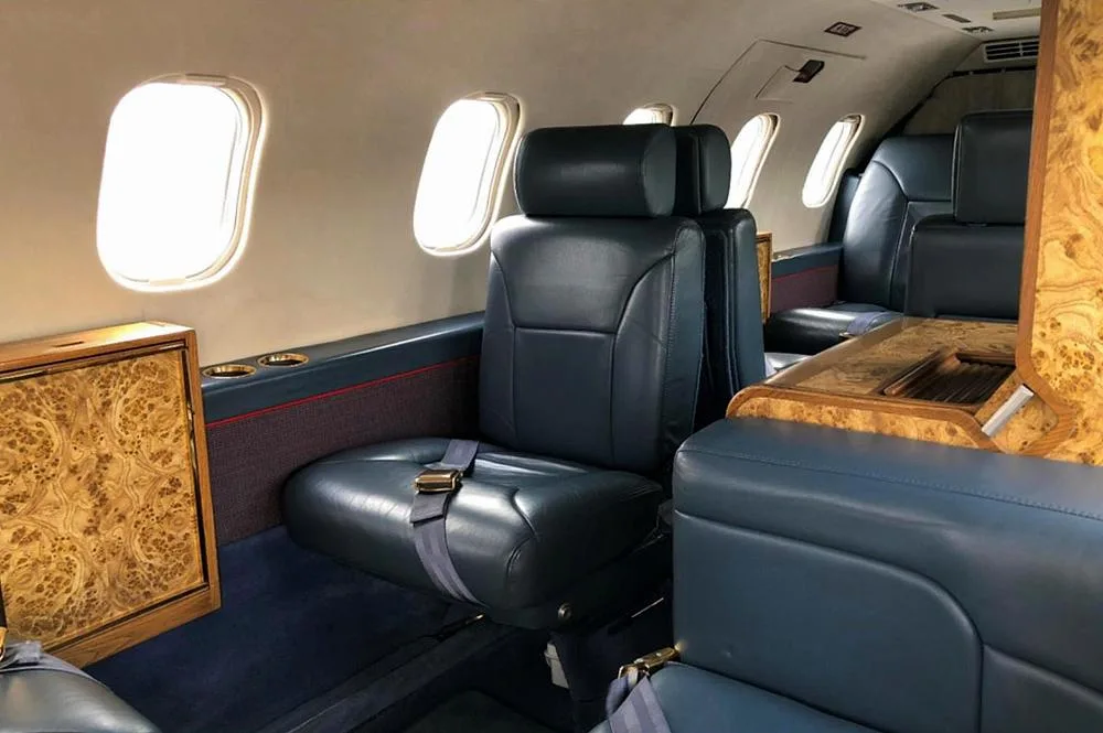 View inside a private jet cabin showing dark blue leather seats, large windows, and wood paneling.