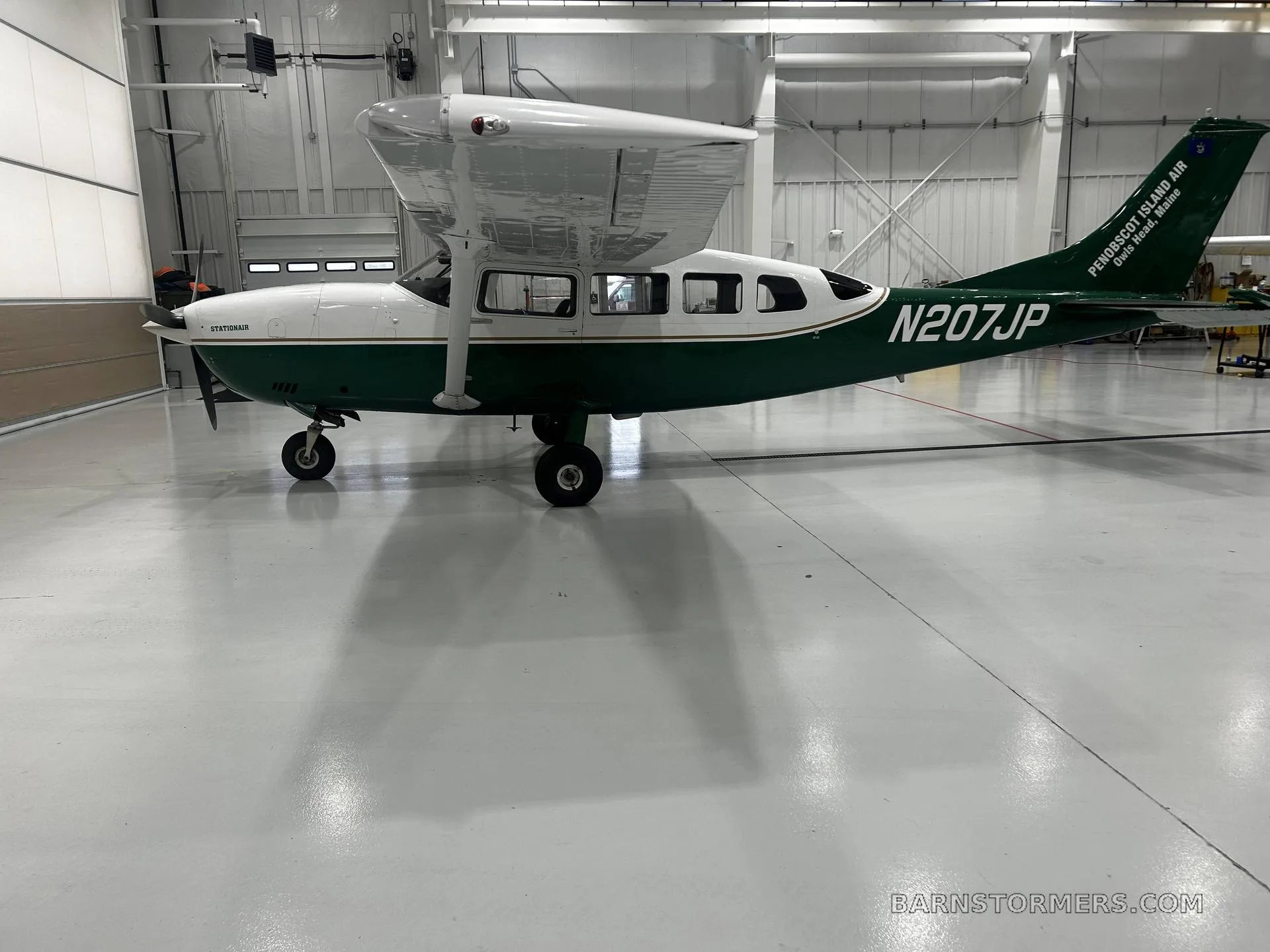 A white and green Cessna 207 Stationair, registration N207JP, is parked in a hangar, viewed from the right side.