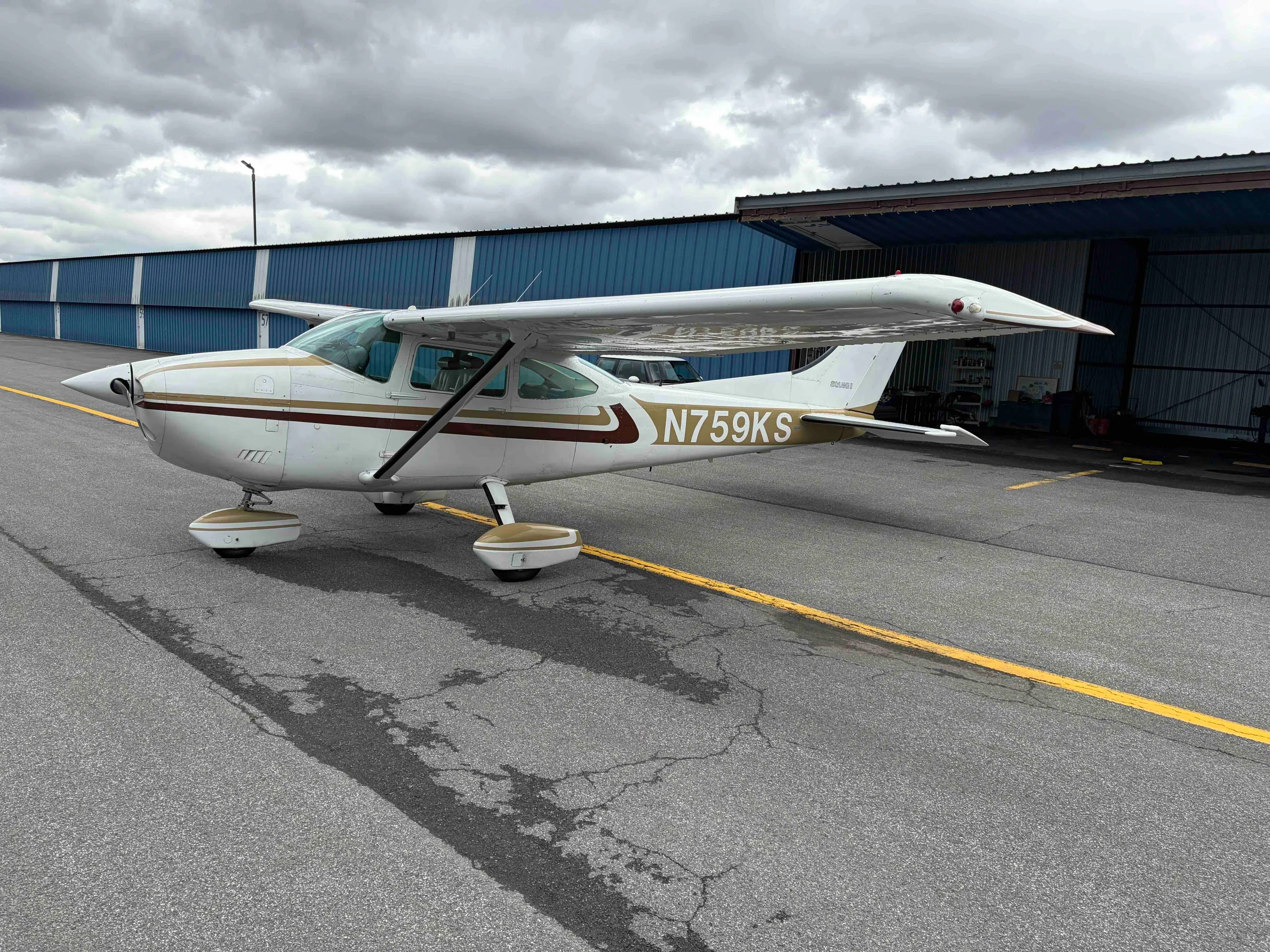 A white Cessna 182 Skylane with gold and brown stripes, registration N759KS, parked on an asphalt tarmac next to blue hangars.