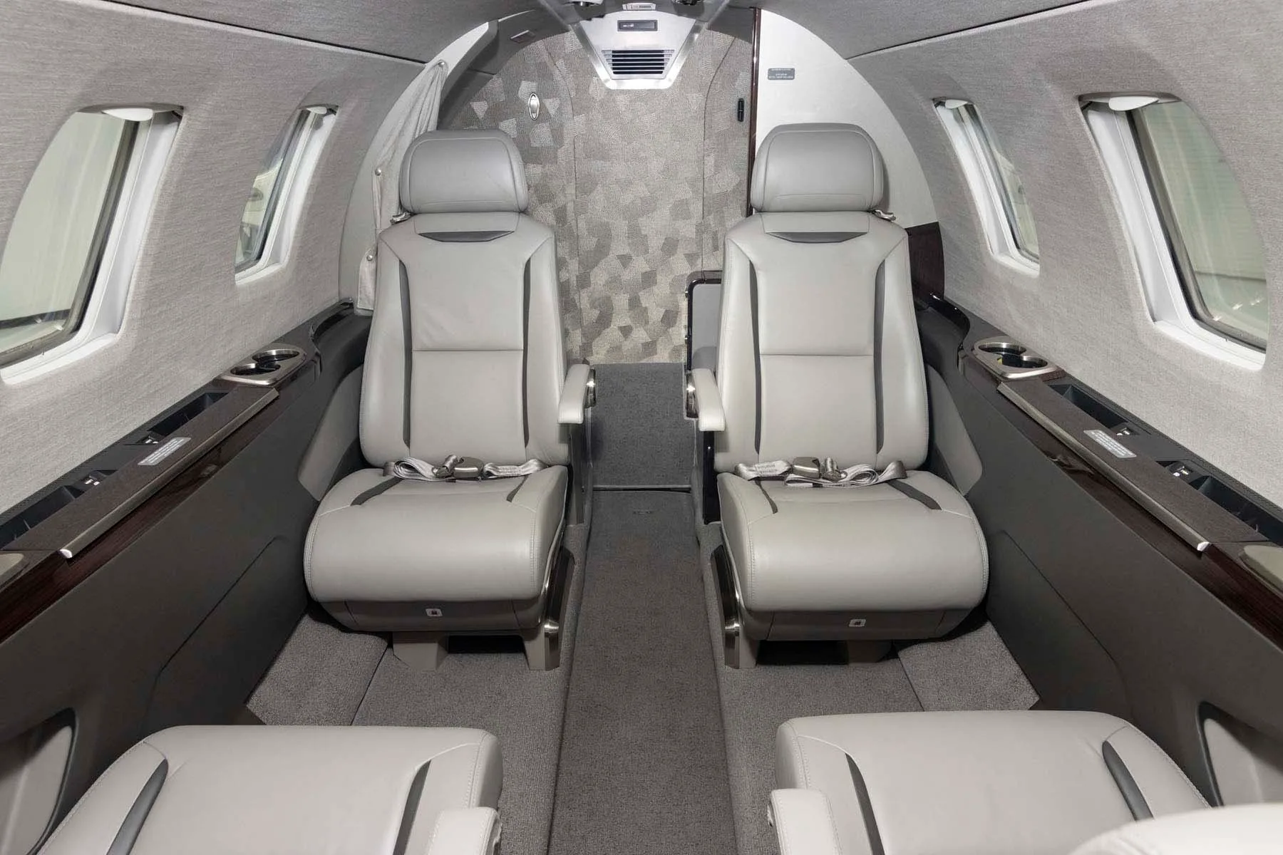 The interior cabin of a private jet, showing two grey leather passenger seats and a patterned wall.
