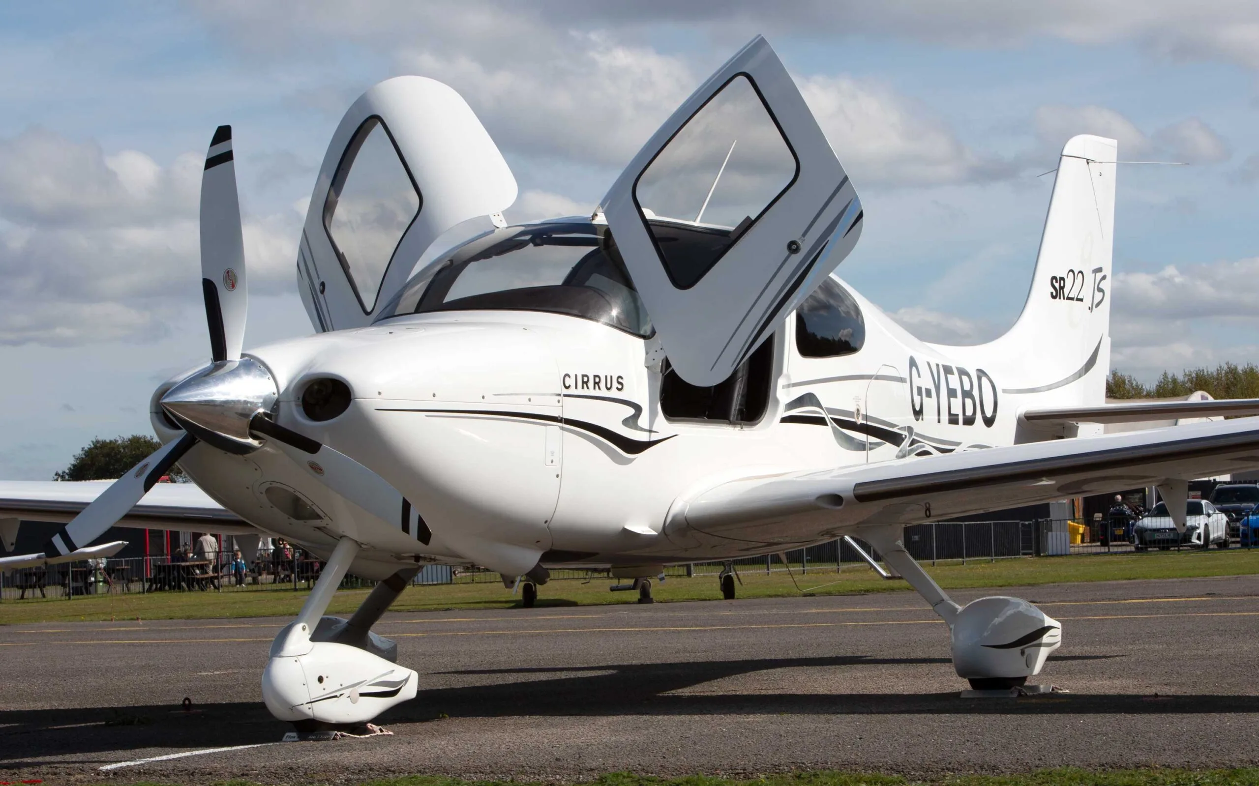 A white Cirrus SR22 TSi aircraft with black and grey stripes, registration G-YEBO, parked on an airfield with its gull-wing doors open.