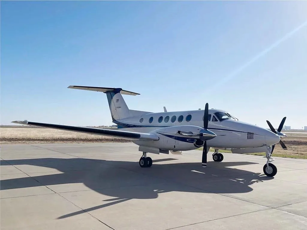 A white and blue Beechcraft King Air twin-turboprop aircraft parked on an asphalt ramp under a clear sky.