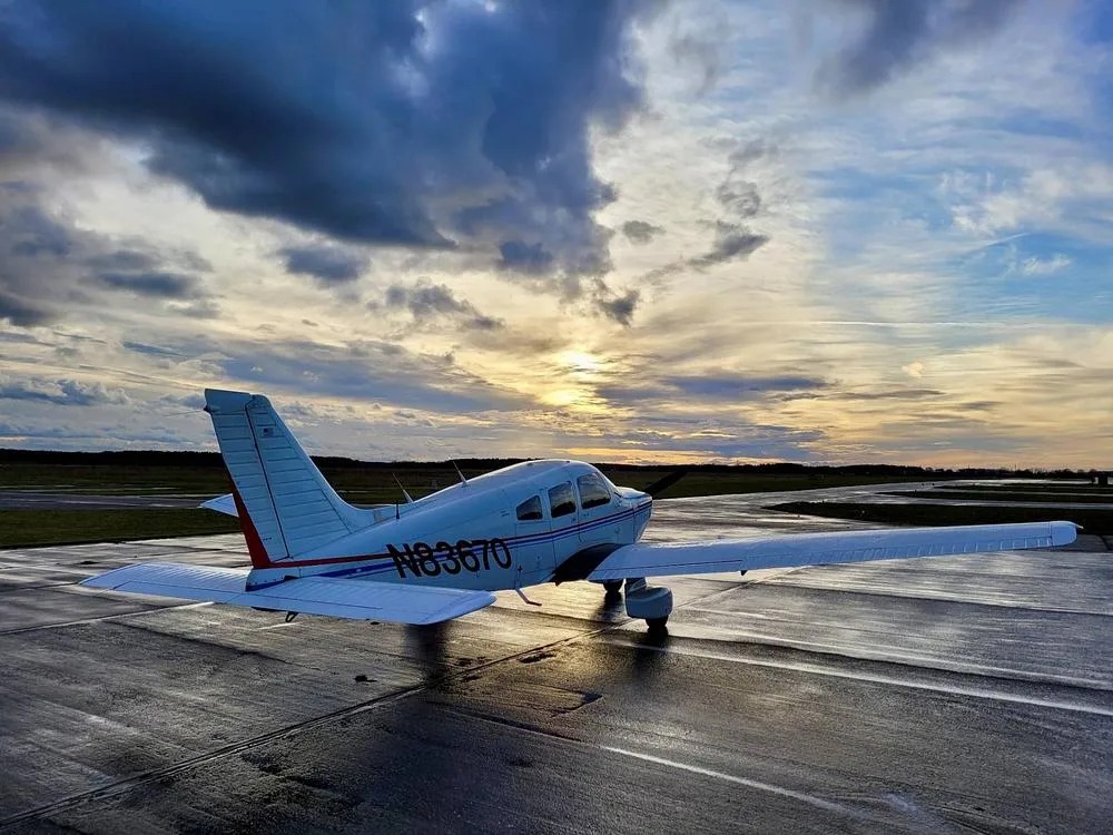 A white Piper Archer II aircraft with red and blue stripes on a wet tarmac at sunset.
