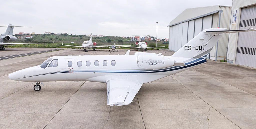 A white Cessna Citation CJ2+ business jet with blue stripes and registration CS-DQT is parked on a tarmac next to a hangar.