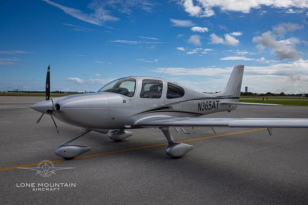 A silver Cirrus SR22 aircraft with black and white stripes on the fuselage, parked on an airfield under a partly cloudy sky.