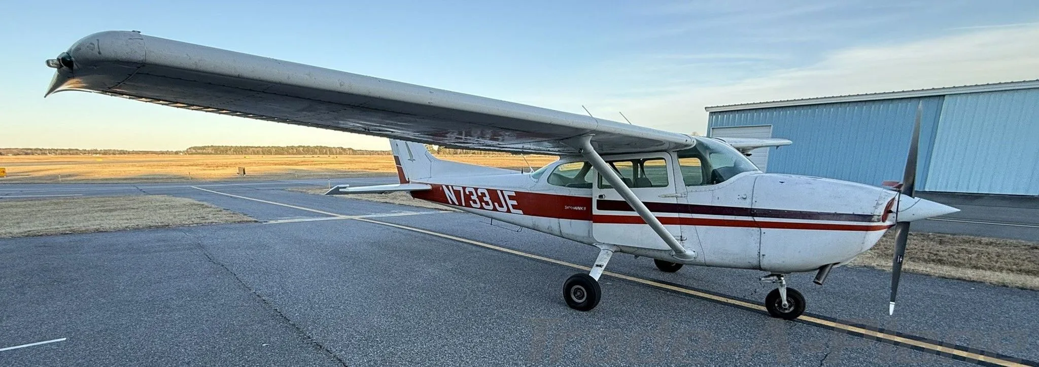 A white Cessna 172N with red and black stripes, registration N733JE, parked on an airfield with hangars in the background.