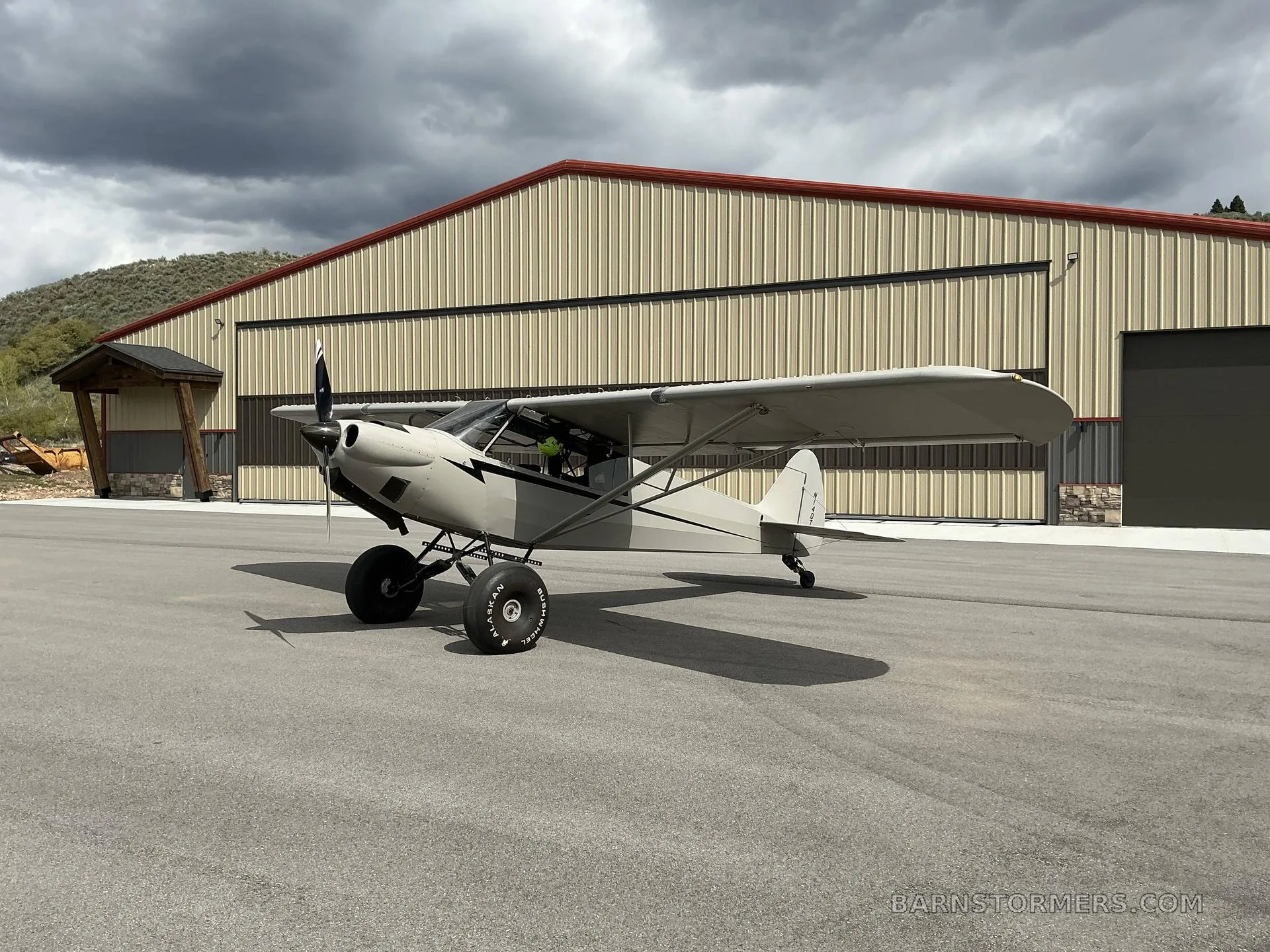 A light grey high-wing taildragger aircraft with black stripes and large tundra tires, parked on asphalt in front of a hangar with mountains in the background under a dramatic cloudy sky.