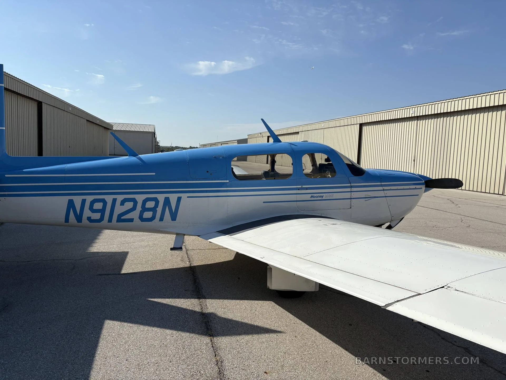 A blue and white Mooney M20 aircraft (N9128N) parked on an airfield next to hangars, viewed from the left side.