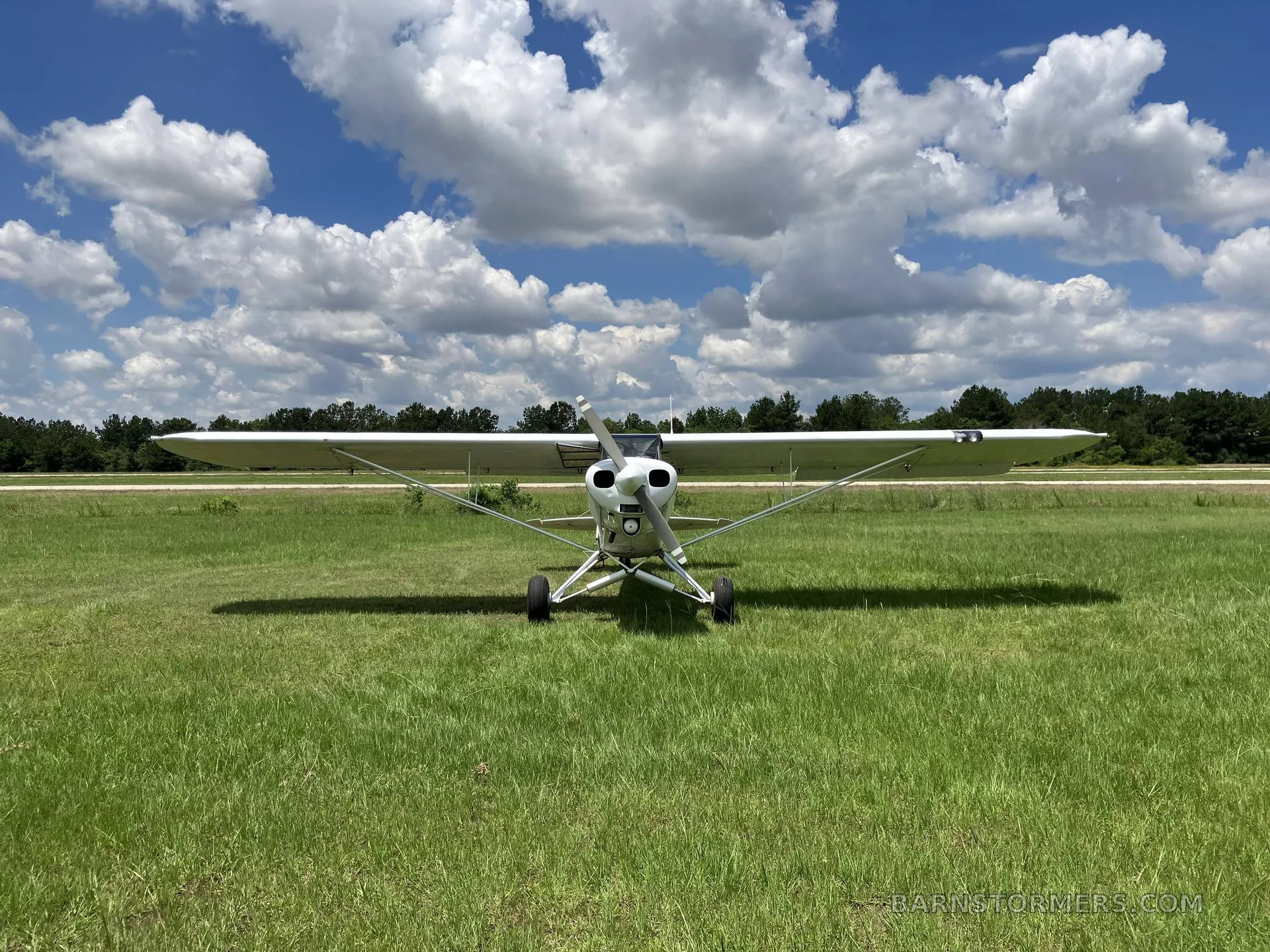 A white single-engine high-wing aircraft with a propeller, viewed from the front, on a grassy field under a blue sky.