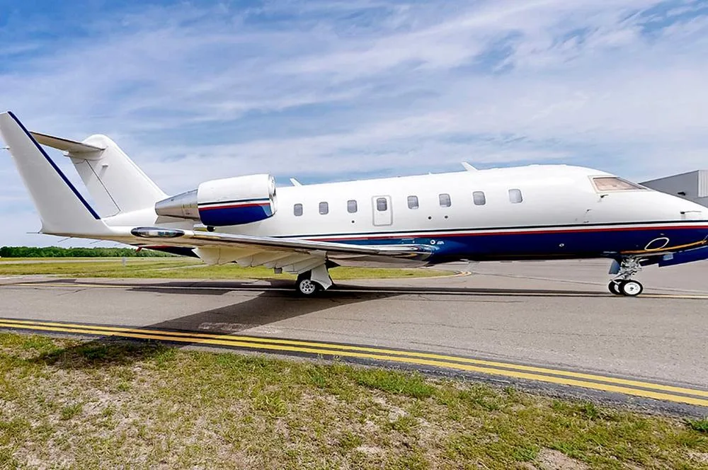 A white and blue Bombardier Challenger 650 business jet is parked on an airport tarmac under a partly cloudy sky.