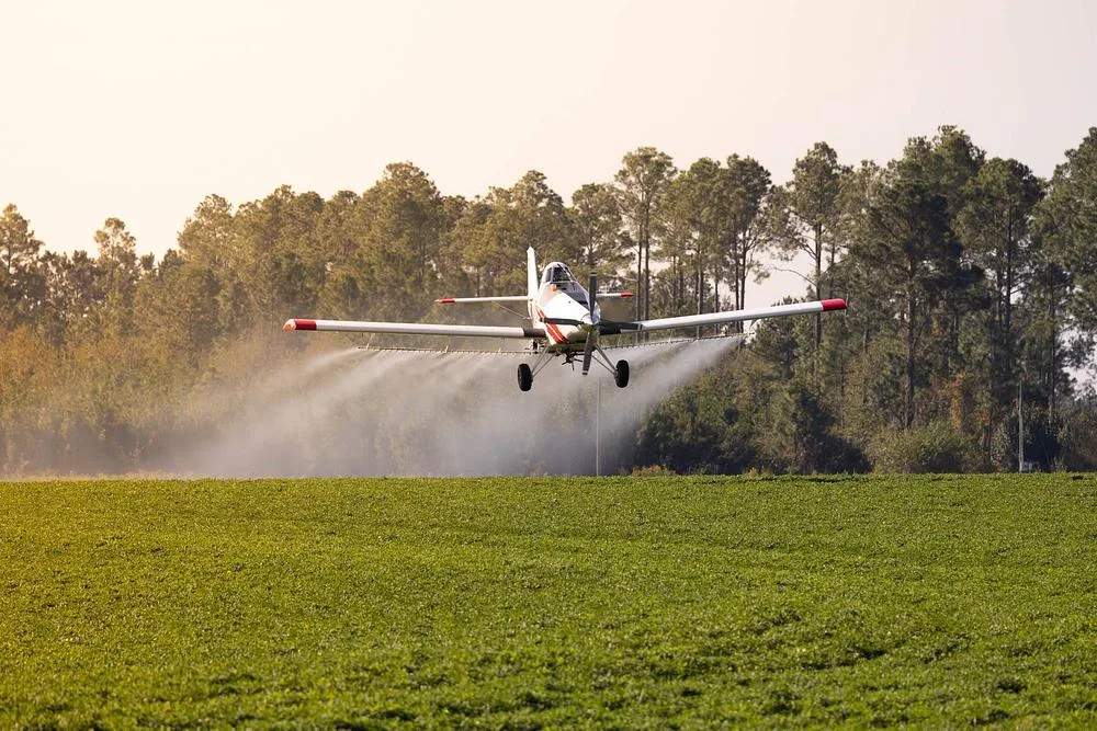 A white and red crop duster aircraft spraying a green field with trees in the background.