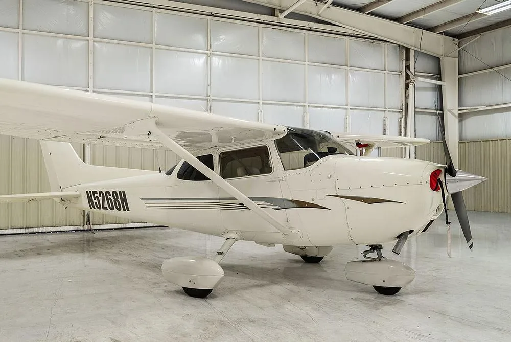 A side profile of the white Cessna 172, registration N5268H, with gold and gray stripes, parked inside a hangar.