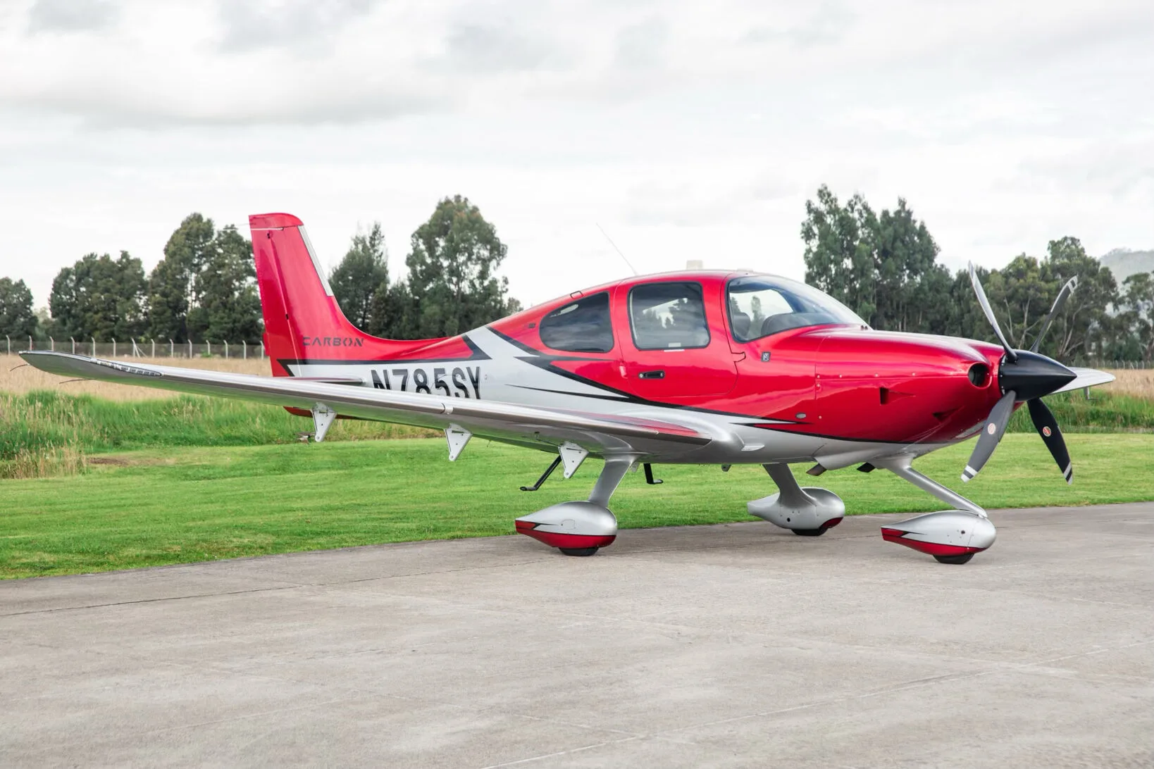 A red and silver Cirrus SR22T aircraft, registration N785SY, parked on an airfield with trees in the background.