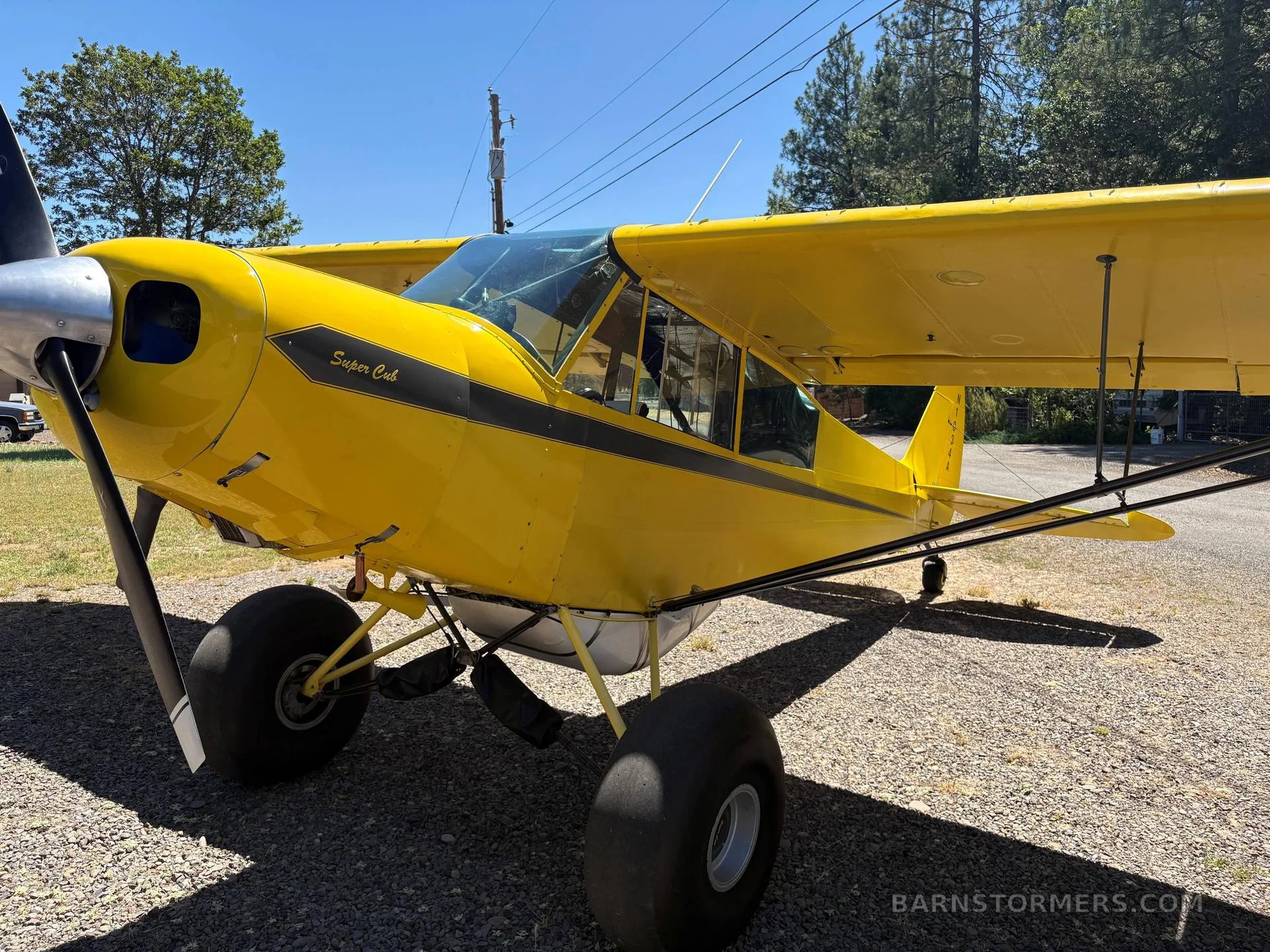 A front-quarter view of a yellow Piper Super Cub with large tundra tires and a black propeller, parked on a gravel surface.