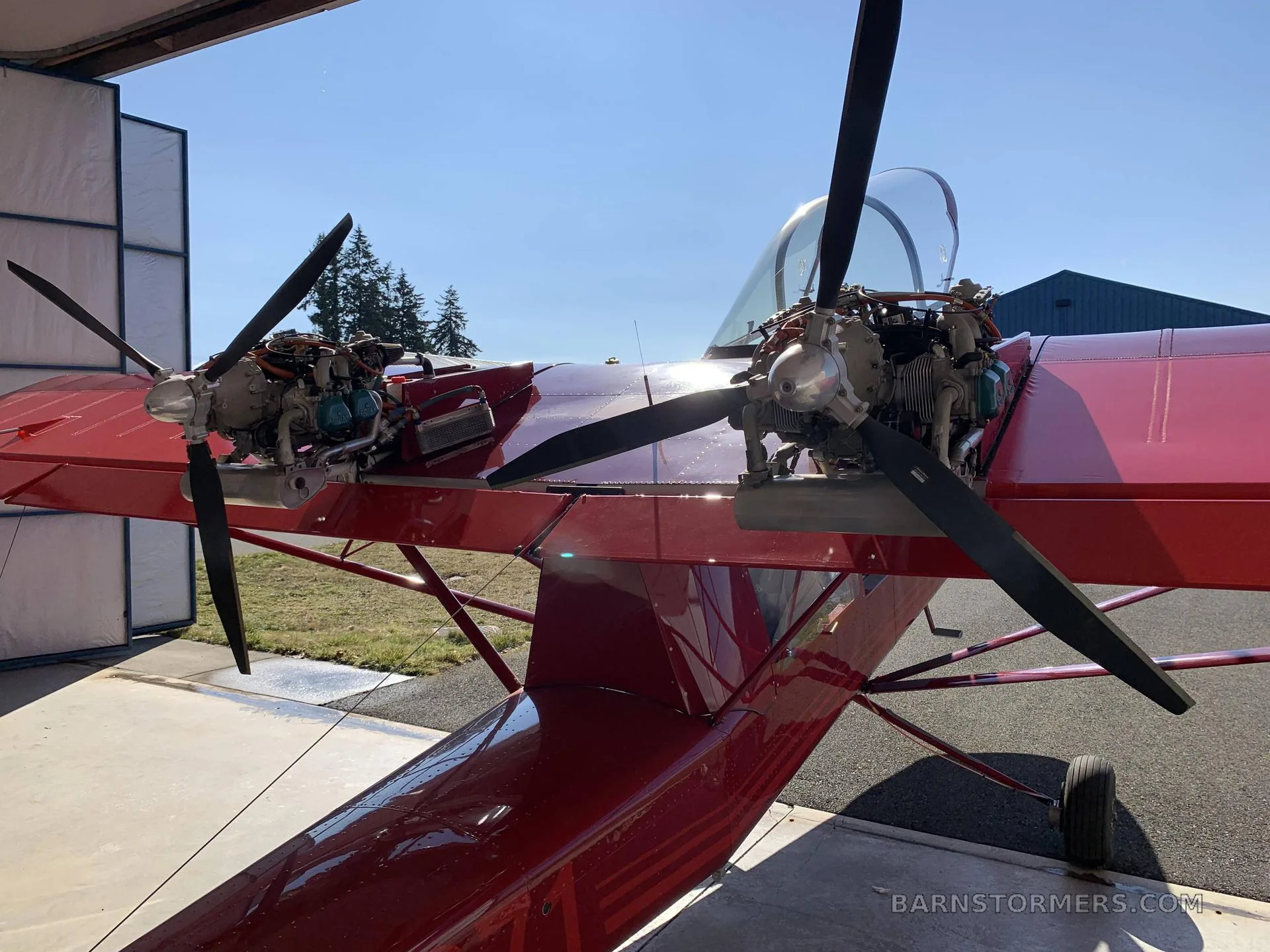 A close-up view of the twin engines and propellers of a red Air Cam aircraft, parked partially inside a hangar.