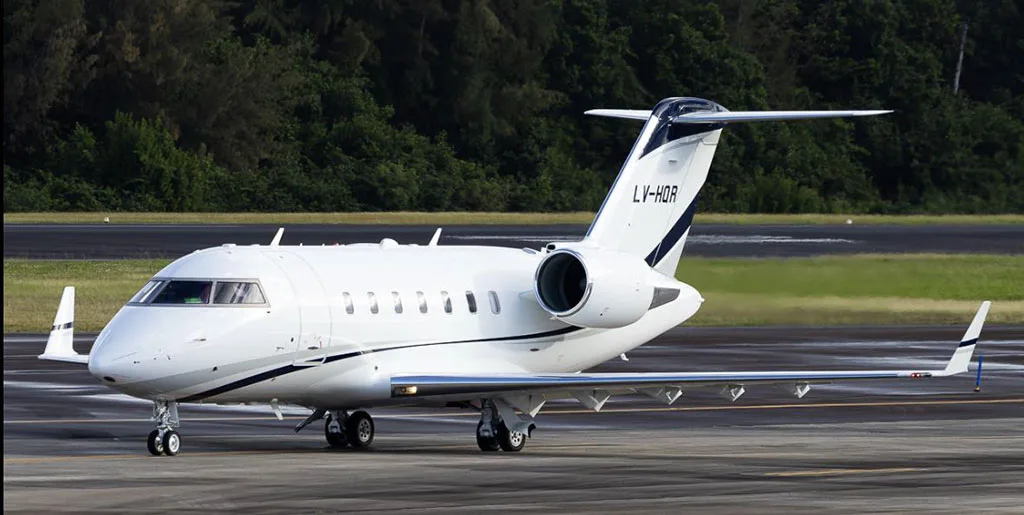 A white business jet with dark blue and grey stripes on the tail and fuselage, registration LV-HQR, is parked on a tarmac with trees in the background.