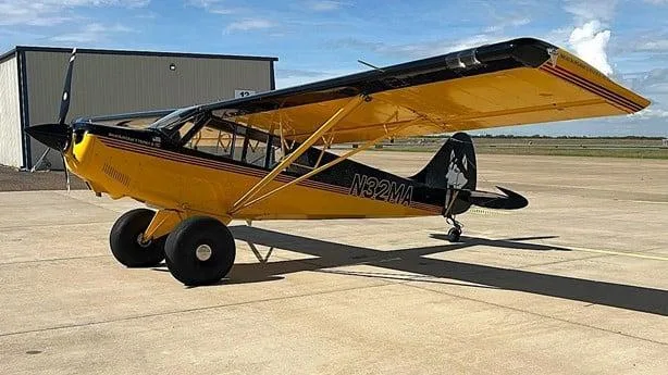 A yellow and black Aviat Husky A-1C aircraft with large tundra tires is parked on a concrete tarmac next to a hangar, viewed from the left side.
