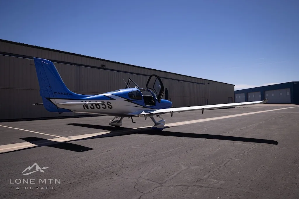 A rear three-quarter view of the blue and silver Cirrus SR22 aircraft, N365S, with its doors open, parked on an asphalt ramp.