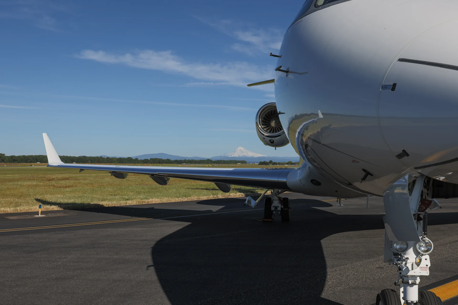 A low-angle view of the front fuselage, wing, and engine of a white business jet on a tarmac, with a distant mountain.