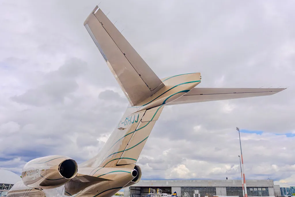 A close-up view of the tail section of a beige business jet with green stripes and registration C-GHJJ against a cloudy sky.