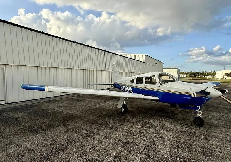 A white and blue Piper Arrow aircraft with registration N338PA parked on an asphalt tarmac next to a white hangar under a cloudy sky.