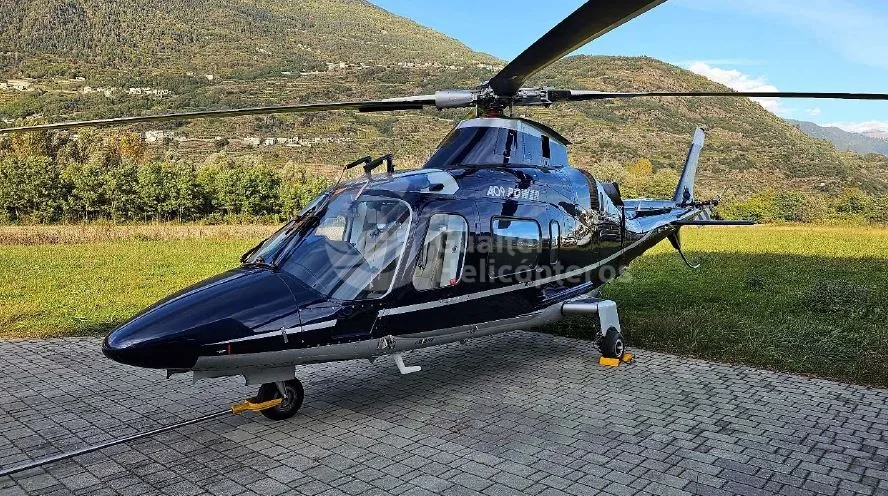 A dark blue Agusta A109 Power helicopter parked on a paved area with mountains in the background.