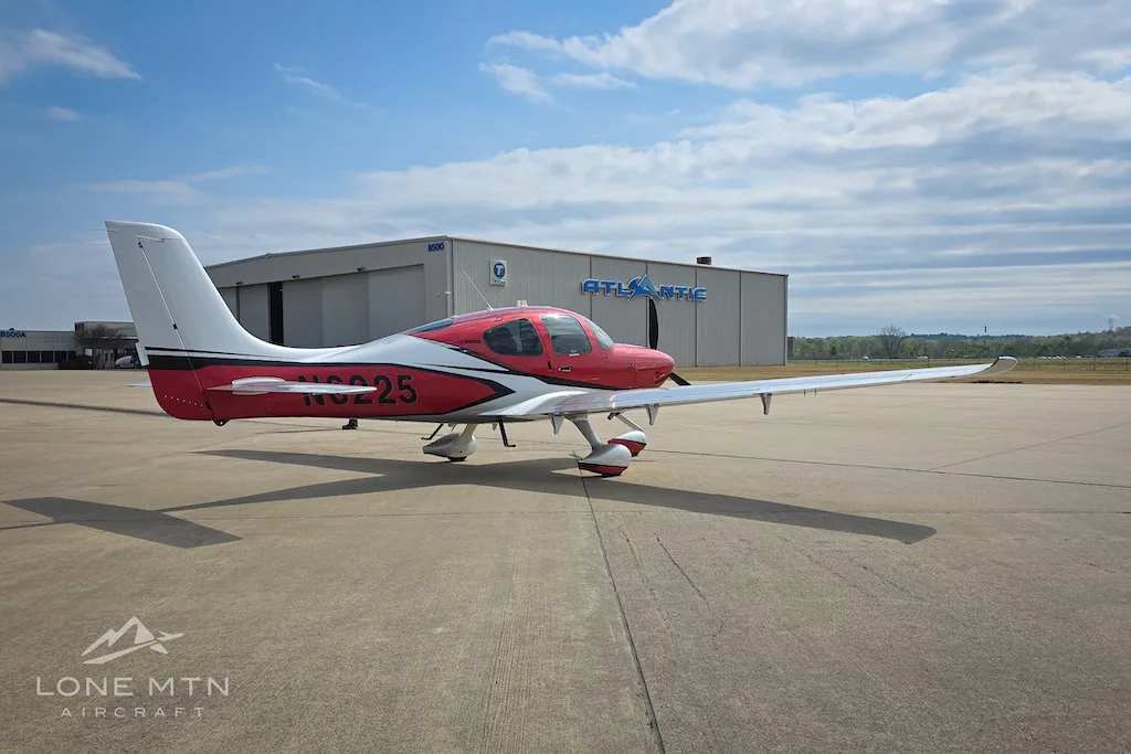 A red and silver Cirrus SR22 aircraft, N6225, parked on an airport apron with a hangar in the background under a blue sky.
