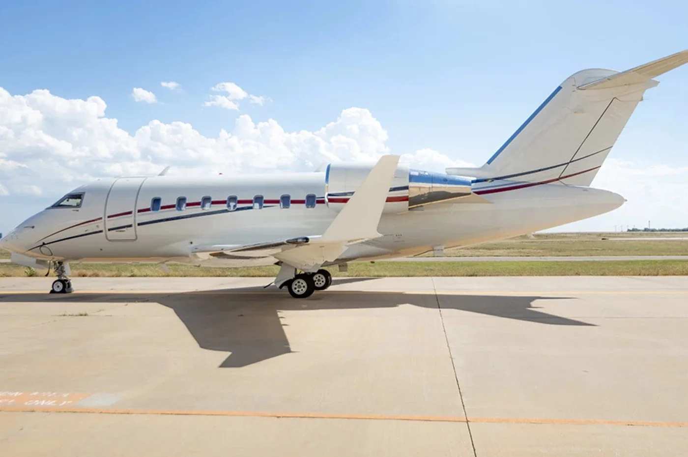 A white Bombardier Challenger business jet with red and blue stripes on the tail and fuselage, parked on an airfield under a partly cloudy sky.