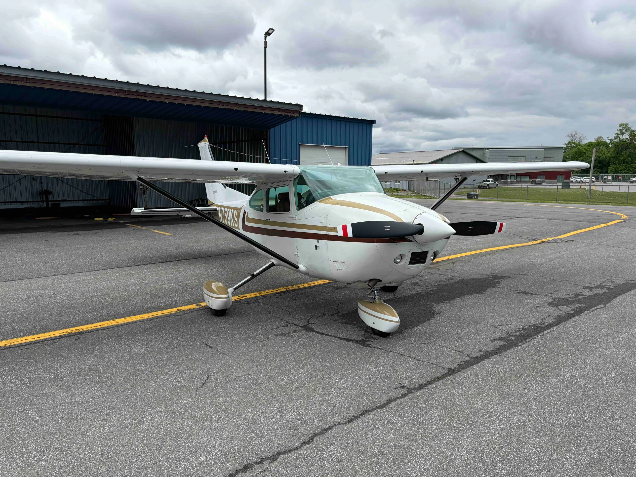 A front view of the white Cessna 182 Skylane N759KS with gold and brown stripes, parked on an airport tarmac.
